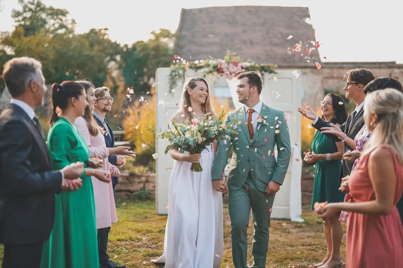 Svatební pár v zelených a bílých šatech zasypaný konfetami hostů na venkovním obřadu.